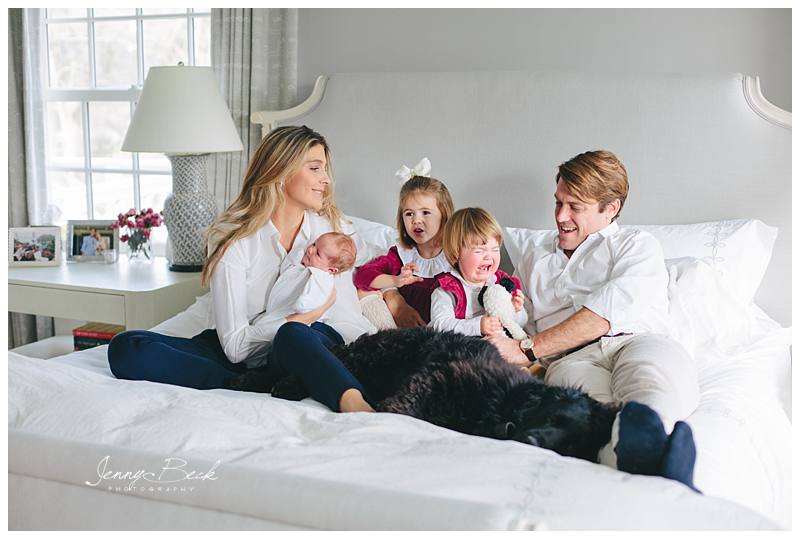 family casually sitting on bed with new baby crying toddler and their dog