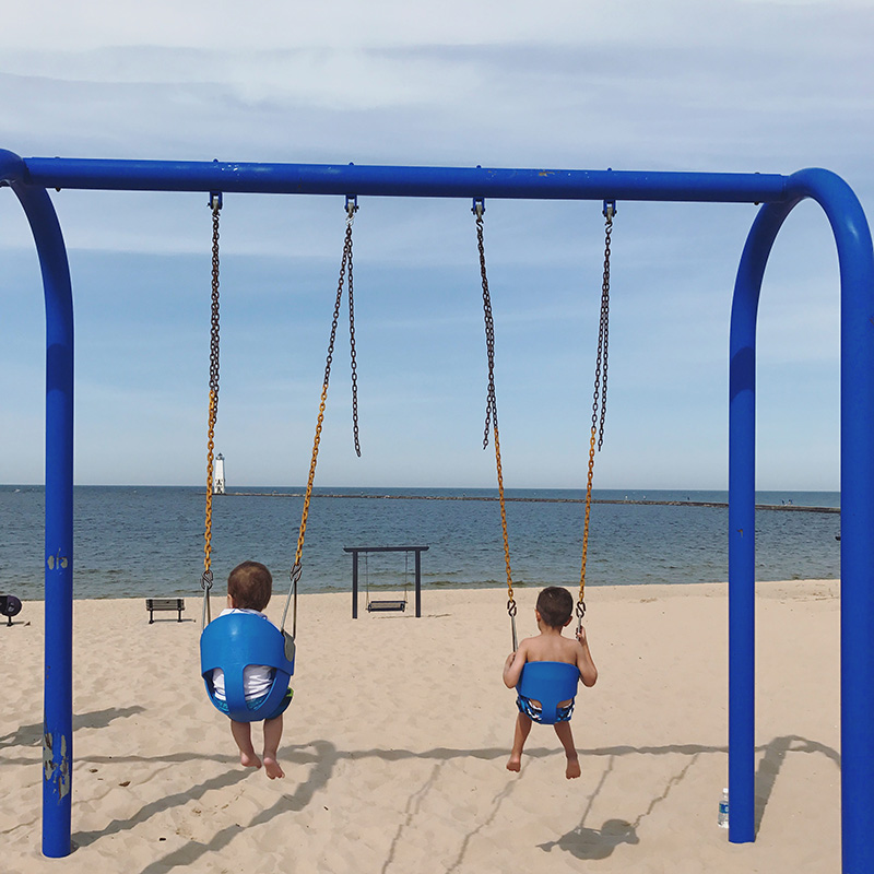 the backside of two young boys swinging on the beach at frankfort michigan