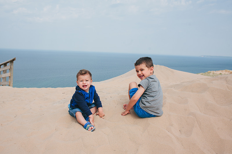 two little boys smiling while playing in the white sand with lake michigan water behind