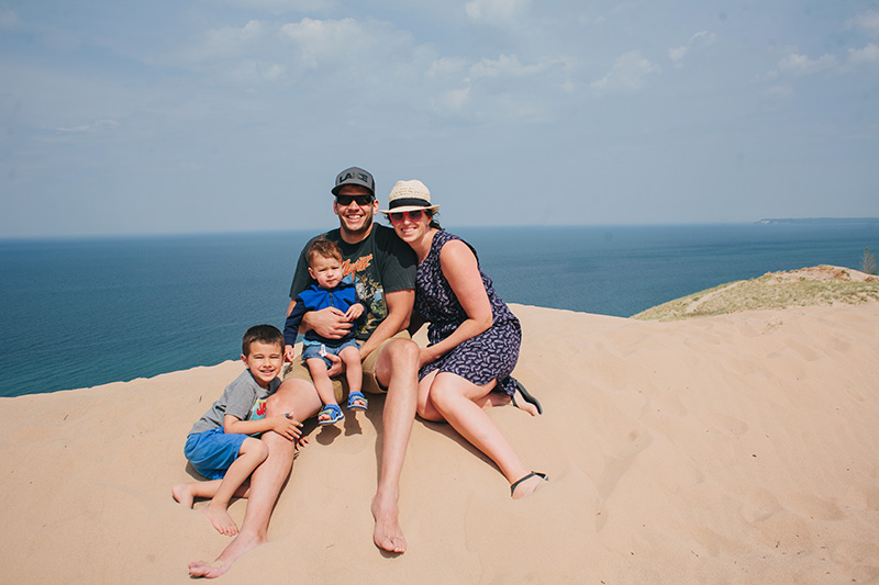 young family sitting in white sand smiling together with turquoise blue lake Michigan water behind.
