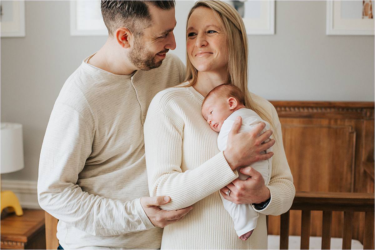 mom and dad looking at each other while mom holds newborn baby wrapped in neutral white wrap